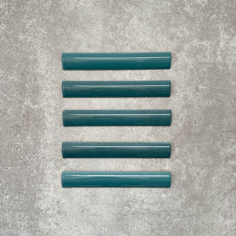 Flatlay von Zellige Pfauengrün Bullnose-Keramikfliesen im marokkanischen Stil im Format 3 × 20 cm, mit abgerundetem Kantenprofil, tief blau-grüner Glasur und feiner tonaler Variation auf neutralem Hintergrund.