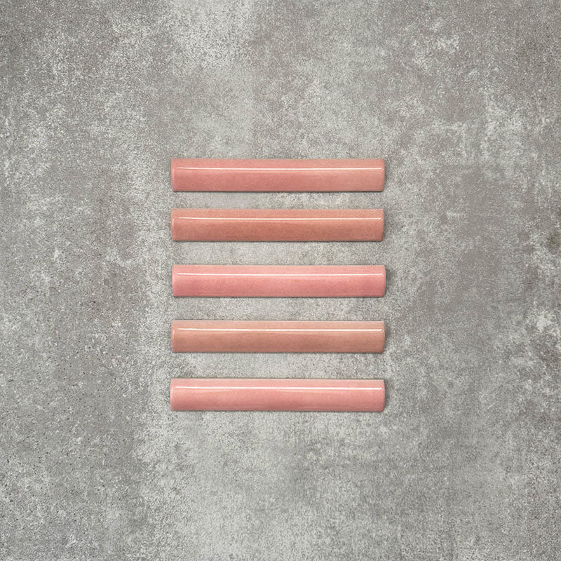 Flatlay mit Zellige Rosa Bordürenfliese im Format 3 × 20 cm im marokkanischen Stil. Keramische Abschlussfliesen in sanften Blush- und Pudertönen mit glatter, abgerundeter Kante und feiner Glasurvariation, arrangiert auf einem strukturierten grauen Hintergrund, um die handgefertigte Zellige-Optik und das dekorative Abschlussprofil hervorzuheben.