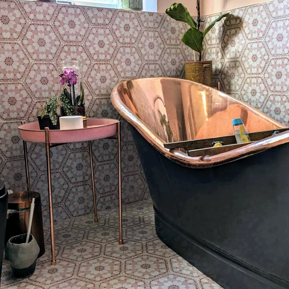 Badezimmer mit Medina Rosa Feinsteinzeug-Hexagonfliesen im marokkanischen Stil im Format 18,7 × 21,6 cm, an Wand und Boden verlegt. Das sanfte geometrische Muster in Rosatönen bildet einen ruhigen Hintergrund und wird mit einer Badewanne in Kupferoptik sowie Vintage-Accessoires stilvoll ergänzt.