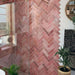 Badezimmerwand mit Zellige Rosa Brick Keramikfliesen im marokkanischen Stil im Format 6,5 × 20 cm, im Fischgrätmuster verlegt. Sanfte Blush- und Pudertöne in Rosa werden mit Wand- und Armaturen in Messing, einer grün gestrichenen Tür und einem Spiegel mit Rundbogen kombiniert – für ein warmes, charaktervolles Finish mit handgefertigter Zellige-Optik.
