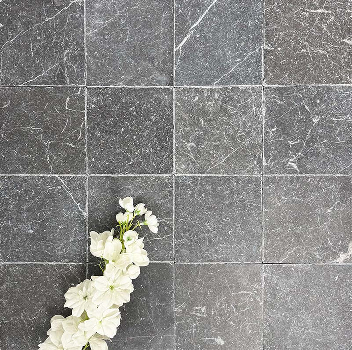 Flatlay mit Fayrouz Black Naturmarmorfliesen im Format 15,2 × 15,2 cm mit getrommelter Oberfläche. Tiefe Anthrazittöne und natürliche Maserungen werden durch weiche weiße Blumen stilvoll ergänzt und schaffen Kontrast sowie zusätzliche Textur.