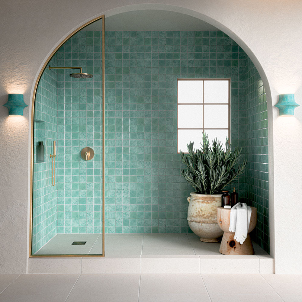 Modern begehbare Dusche mit Bora Bora Aqua Square Flux-Glasur-Wandfliesen im Format 10 × 10 cm, kombiniert mit weißen Bodenfliesen, Duscharmaturen in Messingoptik und einer bogenförmigen Wandnische für ein helles, mediterran inspiriertes Badezimmer.