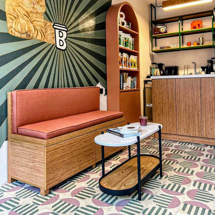 Café-Interieur mit Abstrakter Serit Musterfliese im Format 20 × 20 cm, verlegt als markantes geometrisches Bodendesign. Das verspielte Muster kombiniert sanft rosafarbene Halbkreise mit tiefgrünen Streifendetails auf weißem Grund und wird durch warme Holzmöbel und rosafarbene Sitzmöbel ergänzt – für einen zeitgemäßen, charaktervollen Gewerberaum.