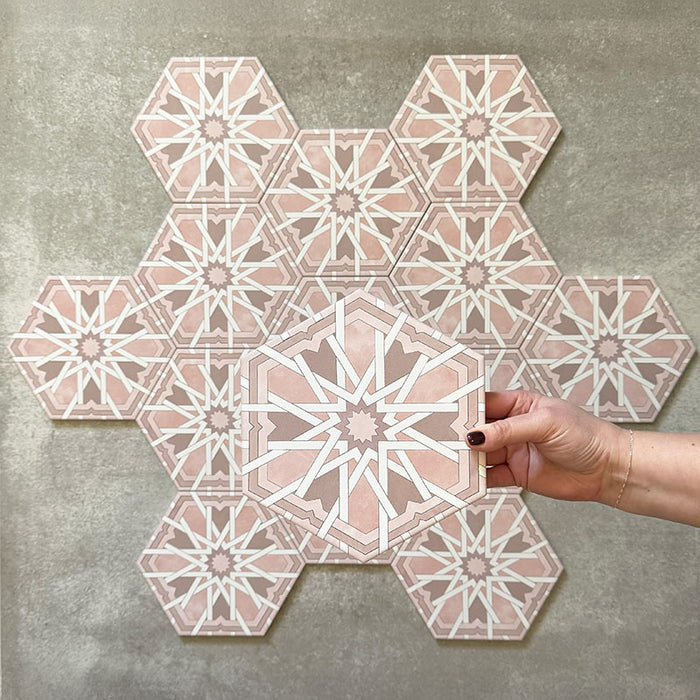 Hand, die eine Medina Hormuz Rosa sechseckige Porzellanfliese im marokkanischen Stil (18,7 × 21,6 cm) hält, vor einem Gruppendesign, um das markante sterninspirierten geometrische Muster in sanften Rosé- und Weißtönen hervorzuheben.