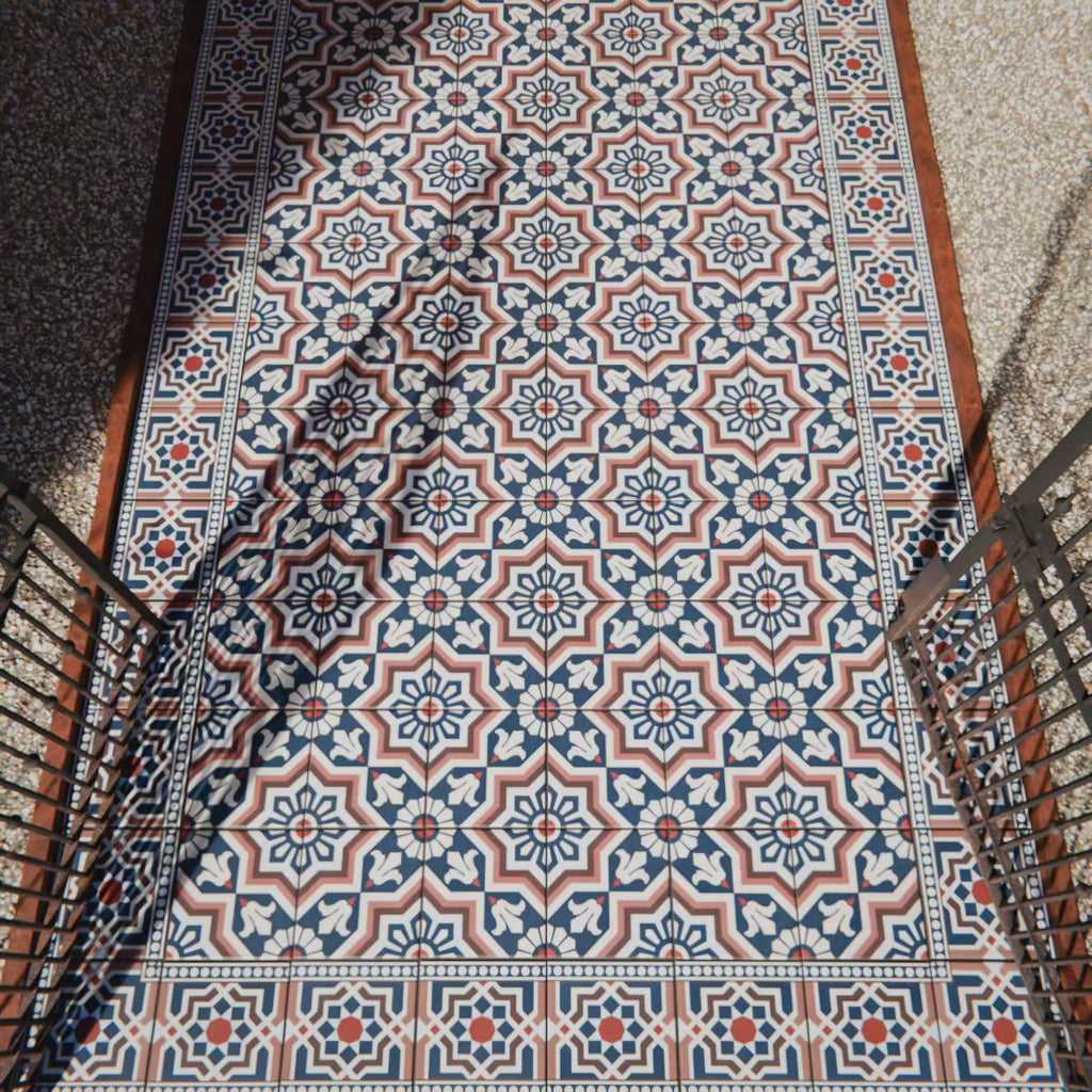 Außenbereich mit Berlin Großartig Bodenfliesen im Format 20 × 20 cm im viktorianisch-marokkanisch inspirierten Stil. Das feine geometrische Muster in Marineblau, Terrakotta und Weiß wird durch schwarze Geländer und natürliches Sonnenlicht stilvoll ergänzt.
